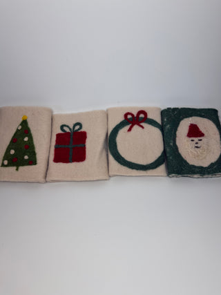 Four square cream-colored felt coasters with Christmas-themed designs, including a green Christmas tree, a red gift box, a green wreath with a red bow, and a Santa Claus face on a dark green background, arranged in a row on a white surface.